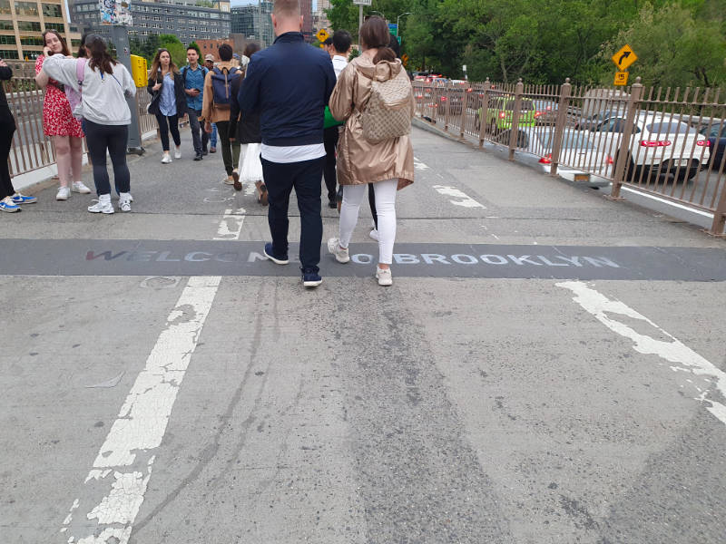 Personas de espalda llegando a Brooklyn luego de cruzar caminando el puente. En el pavimento se lee un letrero en bronce que dice "Welcome to Brooklyn" Foto de Andrea Hoare Madrid. Todos los derechos reservados.
