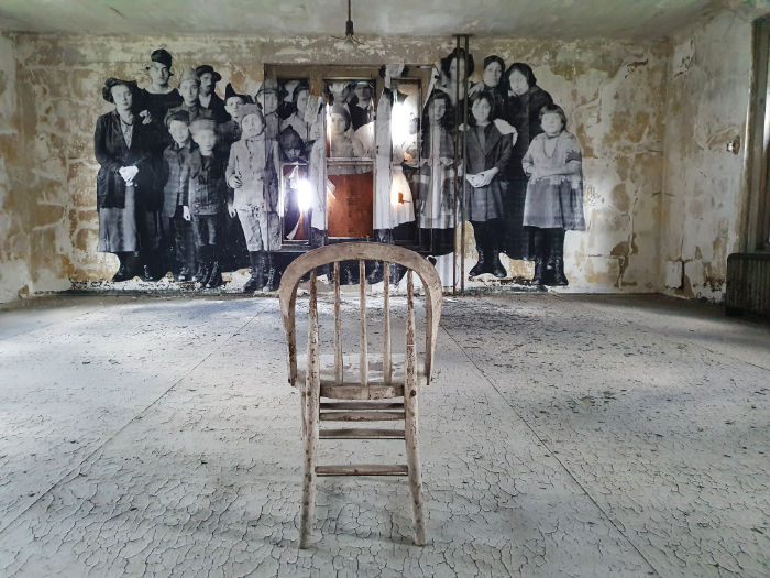 Interior de una de las salas del Hospital de Inmigrantes de Ellis Island. Foto de Andrea Hoare Madrid durante el recorrido del Hard Hat Tour. Se ve una antigua silla vacía en una sala abandonada y sin restaurar, en el fondo una  gigantografía de una foto de un grupo de inmigrantes que pasaron por el Hospital.